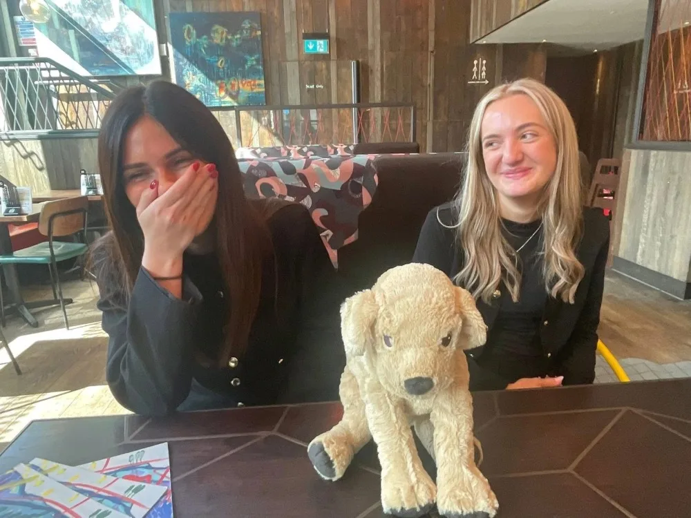 Two people laughing at a dining table with a plush dog on it.
