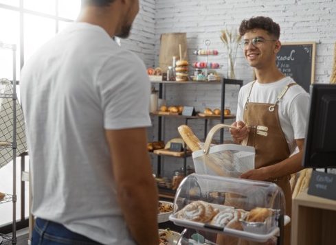 Two men interacting at a bakery counter with one acting as a customer and the other as a baker in a cozy indoor shop setting